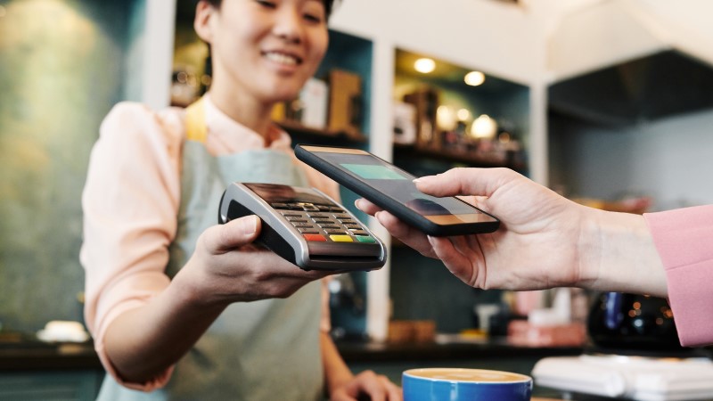 Young female Asian barista holding payment terminal while customer paying for cappuccino with modern smartphone, close-up view